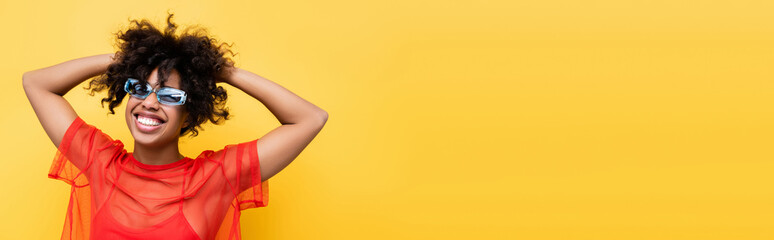 joyful african american woman in red top and blue sunglasses posing isolated on yellow, banner.