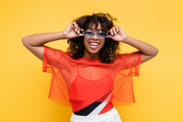 happy african american woman in trendy summer clothes adjusting sunglasses isolated on yellow.