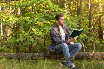 Young man alone resting and reading book in summer green forest outdoors in nature in sunlight