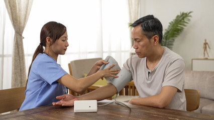 Chinese woman care attendant checking senior man’s blood pressure using a digital sphygmomanometer during home visit. domiciliary care for elderly people concept