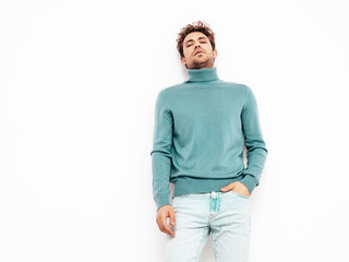 Portrait of handsome confident  model. Sexy stylish man dressed in blue sweater and jeans. Fashion hipster male with curly hairstyle posing near white wall in studio. Isolated