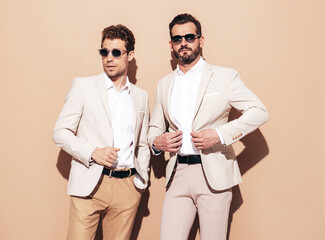 Portrait of two handsome confident stylish hipster lambersexual models. Sexy modern men dressed in white elegant suit. Fashion male posing in studio near beige wall. In sunglasses