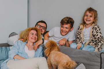  happy friendly family sits at home on the couch, having fun, laughing. Parents play with their children and pets. Dad, teenage son, little daughter, mom, a red poodle. 