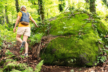 Naklejka na ściany i meble Rear view of young woman in yellow with backpack walking in summer forest next to green moss stone in summer active healthy lifestyle beauty in nature copy space