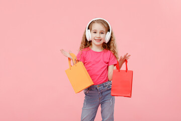 Naklejka premium adorable child girl in headphones is holding shopping bags in hands above head isolated on pink background. shopping concept