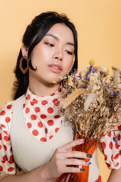 Portrait of stylish asian woman in retro clothes looking at flowers isolated on orange.