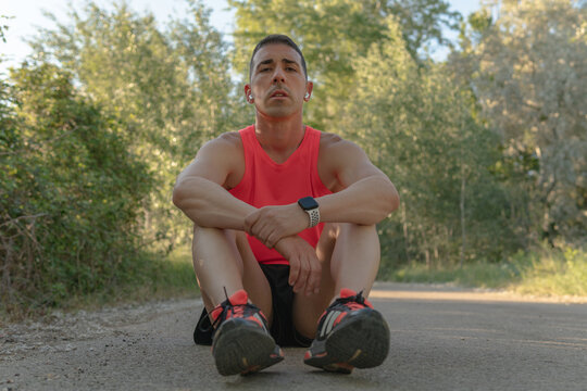 Runner man sitting on asphalt in relaxation position. Front image of a young sportsman in orange sneakers and t-shirt and white digital watch looking at the camera with his hands crossed resting.