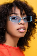 portrait of pretty african american woman in blue and trendy sunglasses isolated on yellow.