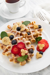 Belgian waffles with strawberries and bananas on a white plate, in the background, a cup of tea and cutlery.