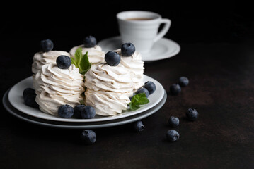 On a dark background, a blurred image of a marshmallow with blueberries on a white plate in the background is a cup of coffee. Concept of coffee drinking.