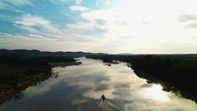 Slow sailing boat on the calm river Ca&ntilde;o Cristales with reflections of the sky at sunset. Around the silhouette of the rainforest, with mountains on the horizon, Colombia