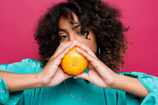 curly african american woman obscuring face with ripe orange isolated on pink.