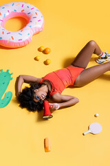 african american woman lying near fruits, swim ring and sunscreen while talking in megaphone on yellow background.