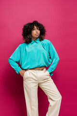 african american woman in turquoise blouse posing with hands in pockets of trousers isolated on pink.