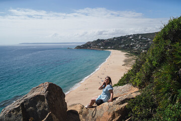 Chica joven en roca observando paisaje de playa con gente realizando parapente delante suya