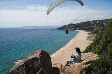 Chica joven en roca observando paisaje de playa con gente realizando parapente delante suya