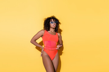 african american woman in sunglasses and swimsuit standing with hand on hip and pointing with finger on yellow background.