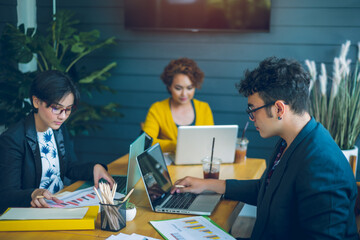 Confident young businessman sitting at office desk with group of colleagues in office They work by using laptop computers and graphing charts to analyze and plan their business.