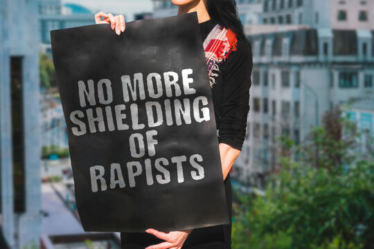 The phrase " No more shielding of rapists " is on a banner in women's hand. A woman holds black cardboard with an inscription. Female. Fight. Help. Crying. Hurt. Justice. Horror. Messy. Gang. Murder