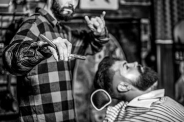 Men barber shaves his beard. Bearded male sitting in an armchair in a barber shop while hairdresser...
