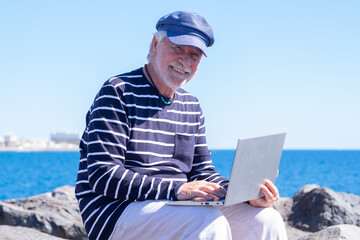 Senior caucasian man sitting on the rocks at sea using laptop in remote working, white-haired elderly male browsing with computer