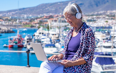 Fototapeta na wymiar Senior caucasian woman with headphones sitting outdoors at sea harbor using laptop in remote working, gray-haired elderly female browsing with computer