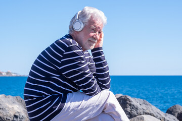 Fototapeta premium Caucasian elderly man with headphones sitting on the cliffs enjoying sea and freedom in vacation