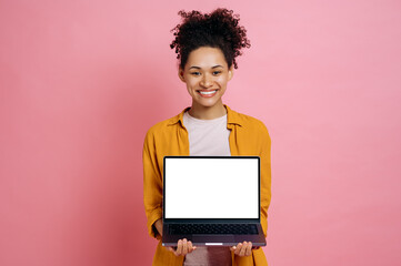 Positive african american girl in casual clothes, holds an open laptop with blank white mock-up screen for advertisement or presentation, stands on isolated pink background, looks at camera, smiles