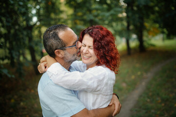 Fototapeta premium slender man with beard kisses and hugs pretty red-haired curly woman. Cute middle aged european couple hugging in the park