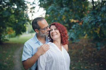 slender man with beard kisses and hugs pretty red-haired curly woman. Cute middle aged european couple hugging in the park