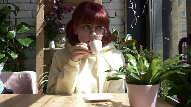 Stylish redhead teenager girl with cup of coffee sit at table in cafe with plants around