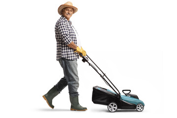 Full length profile shot of a mature man with a straw hat and boots using a lawn mower - obrazy, fototapety, plakaty