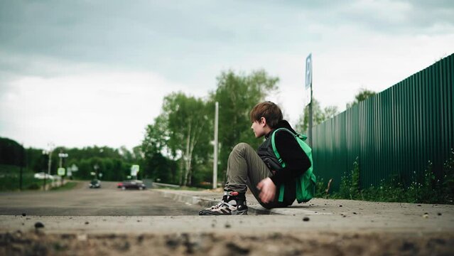 The boy sits down to rest after roller skating on the edge of the road. Shooting from the side