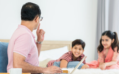 Fototapeta premium Indian father working at home or apartment, raising hand to flaw, warn and ask for silence or quiet with blur background of children playing on bed. Family, Lifestyle, Business, Technology Concept.