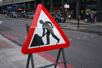 Red triangle warning roadworks sign placed at pavement, blurred city road with people, buses and taxis in background