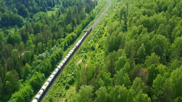 Top, aerial view of long freight train that moving along green forest in summer. Locomotive, tank wagons ride on rails carrying liquid or bulk cargo. Railroad transportation, delivery taken by drone
