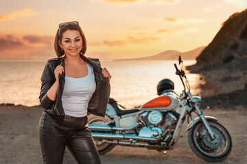 Pretty smiling adult woman in leather jacket posing. Motorbike, sunset and ocean on the background....