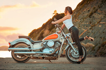 Fototapeta premium Beautiful sexy woman with a high heels and leather pants, posing confidently with motorcycle. Sunset sky on the background. The concept of Motorcyclist Day