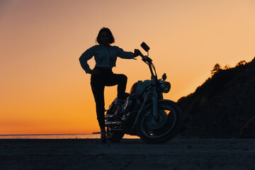 Fototapeta na wymiar Silhouette of sexy woman posing confidently with motorcycle. Orange sunset sky on the background. The concept of Motorcyclist Day
