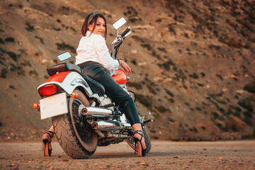 Obraz premium A beautiful adult woman with a high heels and leather pants, sexy posing sitting on motorcycle. Rear view. Rock on the background. The concept of Motorcyclist Day