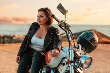 Fototapeta premium Cool smiling adult woman in leather jacket posing near motorcycle. Sunset and ocean on the background. The concept of Motorcyclist Day