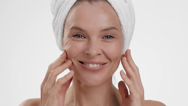 Skin lifting procedures. Close up portrait of happy middle aged lady with towel on head making anti edema facial massage
