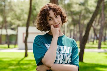 Fototapeta premium Young sad redhead woman wearing green t-shirt standing on city park, outdoors touching chin and looking at the camera, thoughtful, lost in thoughts consider idea. Making important choice.