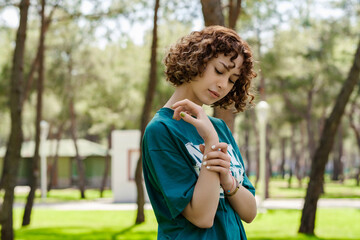 Side view of sad young woman wearing green t-shirt standing on city park, outdoors holding her painful wrist. Suffering pain on hands and fingers, arthritis inflammation.