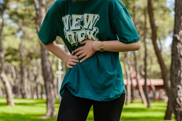 Naklejka premium Cropped view of young sportive woman wearing green t-shirt and black legging standing on city park, outdoors with hand on stomach because indigestion, painful illness feeling unwell. Ache concept.