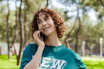 Fototapeta premium Young redhead woman wearing green t-shirt standing on city park, outdoors talking on mobile phone with friends or boyfriend with smiles.