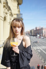 In the afternoon, in the light of the sun, a touching girl in black pajamas enjoys a glass of orange juice. She's good on the balcony.