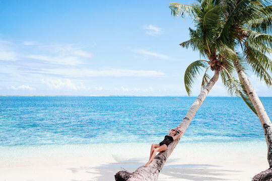 Vacation on the seashore.Young woman in black swimsuit on the beautiful tropical beach laying on the palm tree.