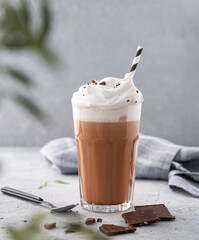 Hot chocolate with whipped cream and chocolate pieces in a tall glass on a blue background with a...