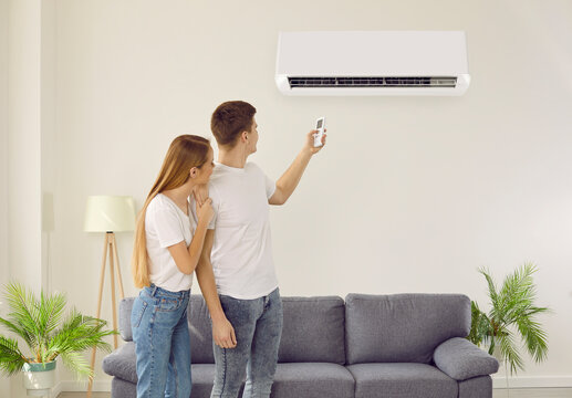 Couple Tenants Turning On Conditioner In Living Room Breathing Fresh Air. Man And Woman Renters Use Condition Device At Home. Indoors Ventilation And Modern Technology.